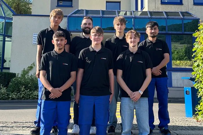 Sieben neue Auszubildende von Linamar Plettenberg in schwarzen Poloshirts stehen in zwei Reihen vor einem Gebäude.