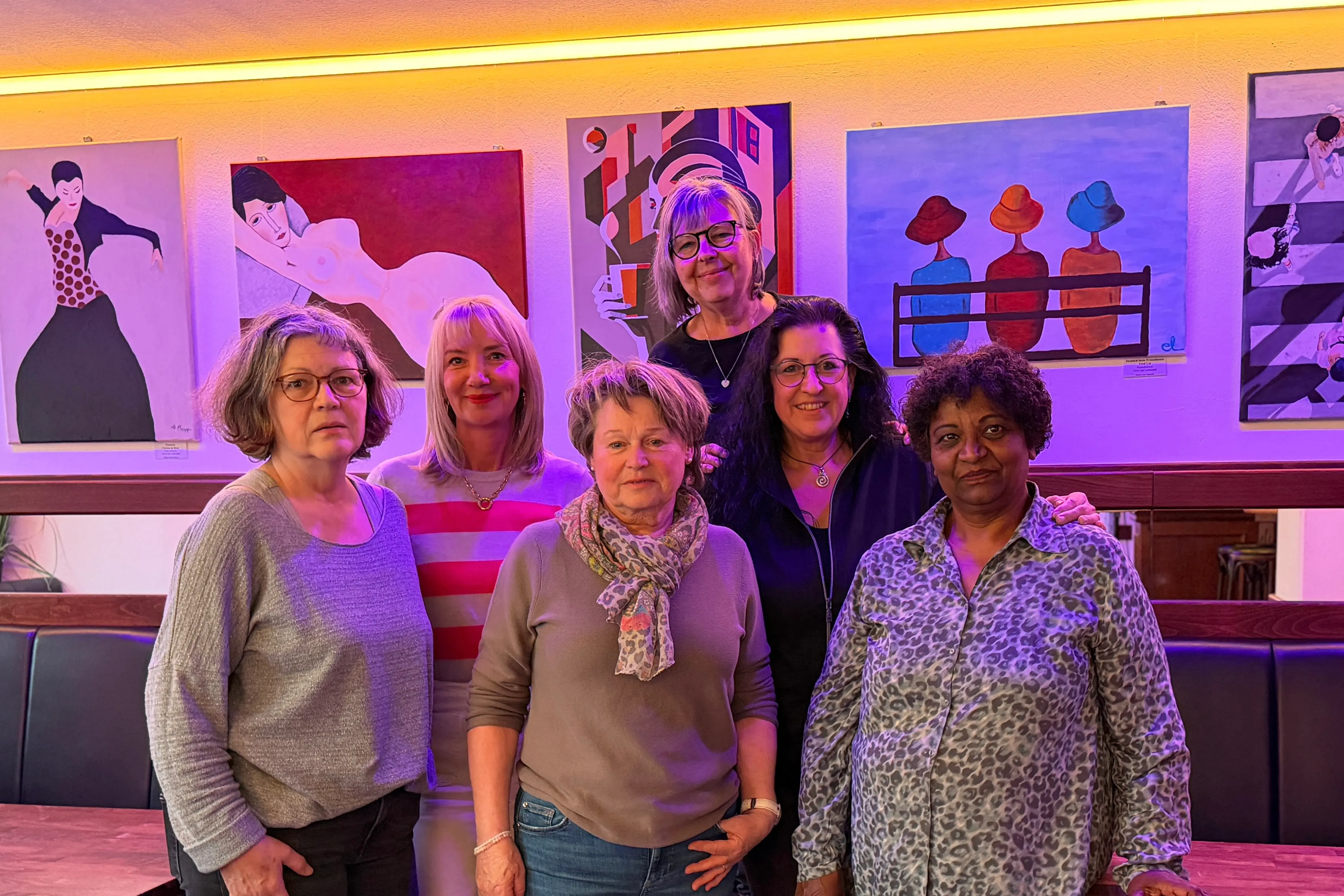 Fünf Frauen stehen vor einer Wand mit abstrakten Gemälden in einem Café. Sie lächeln in die Kamera und zeigen die Ergebnisse ihres Malkurses.