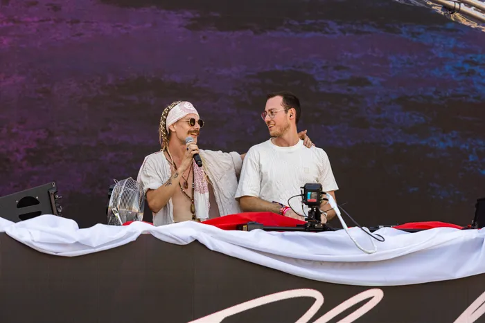 Zwei lächelnde Männer auf einer Bühne beim Ferik Festival; einer trägt ein Kopftuch, eine Sonnenbrille und hält ein Mikrofon.