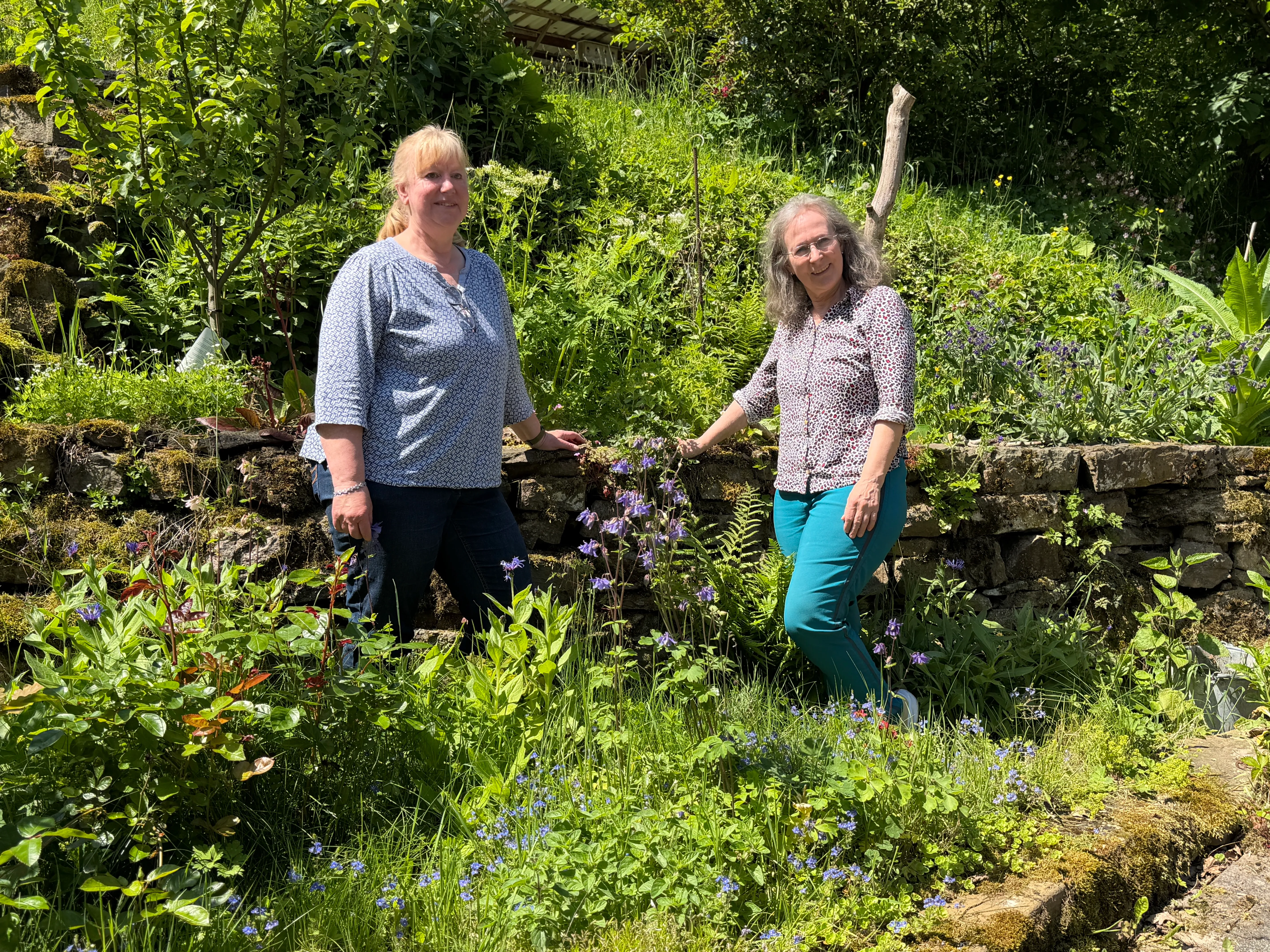 Zwei Frauen stehen in einem üppigen Naturgarten vor einer Steinmauer mit blühenden Pflanzen. Im Vordergrund sind verschiedene Blumen und Kräuter zu sehen.