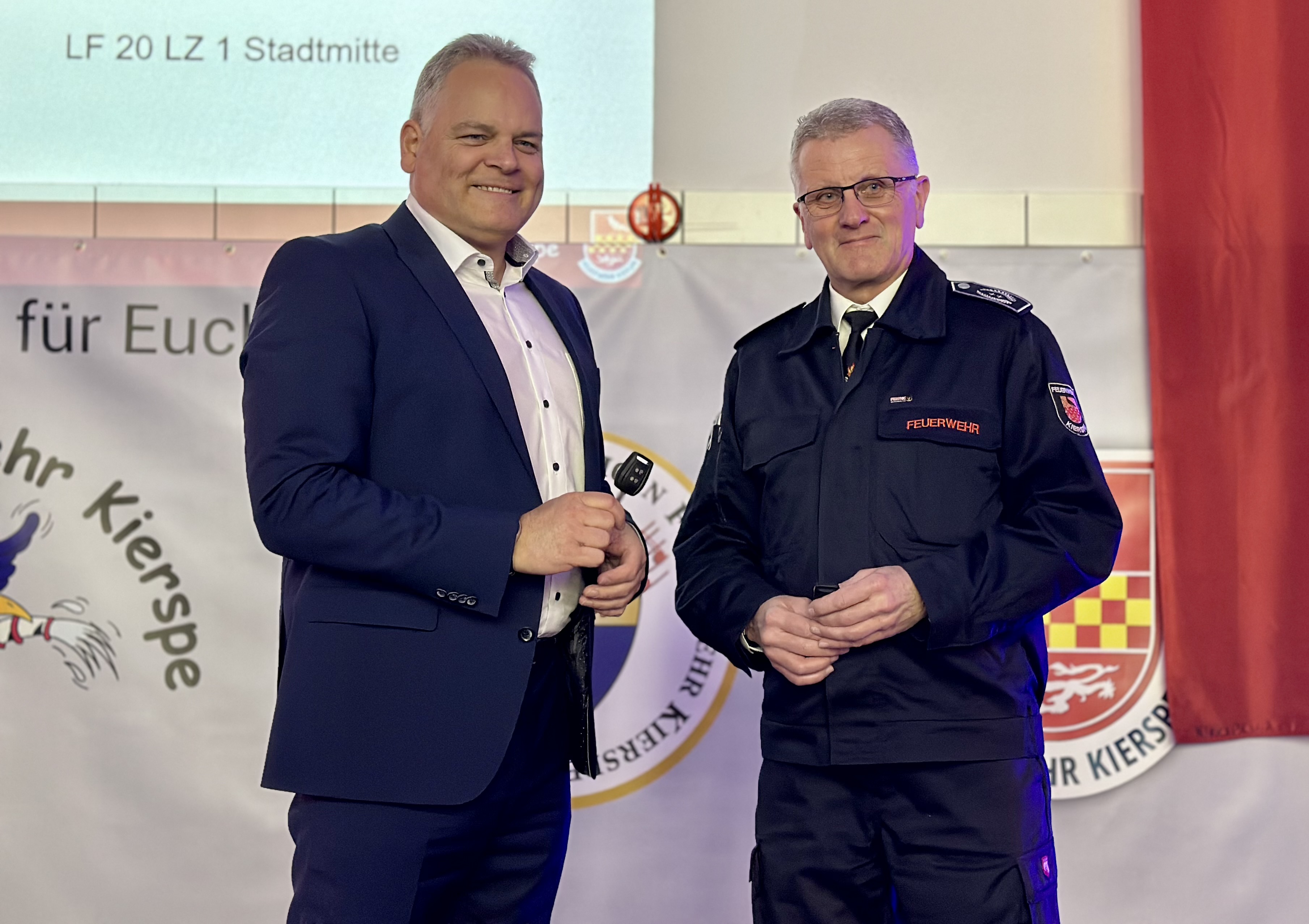 Bürgermeister Olaf Stelse und Feuerwehrleiter Mike Budde stehen auf der Bühne und sprechen in ein Mikrofon. Im Hintergrund ein Banner mit dem Aufdruck „Feuerwehr“ und dem Stadtwappen.