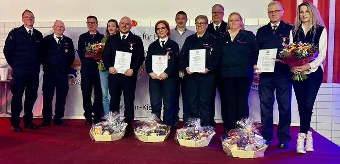 Acht Feuerwehrleute in Uniform stehen nebeneinander auf einer Bühne und halten Urkunden und Blumensträuße. Vor ihnen stehen Geschenkkörbe.