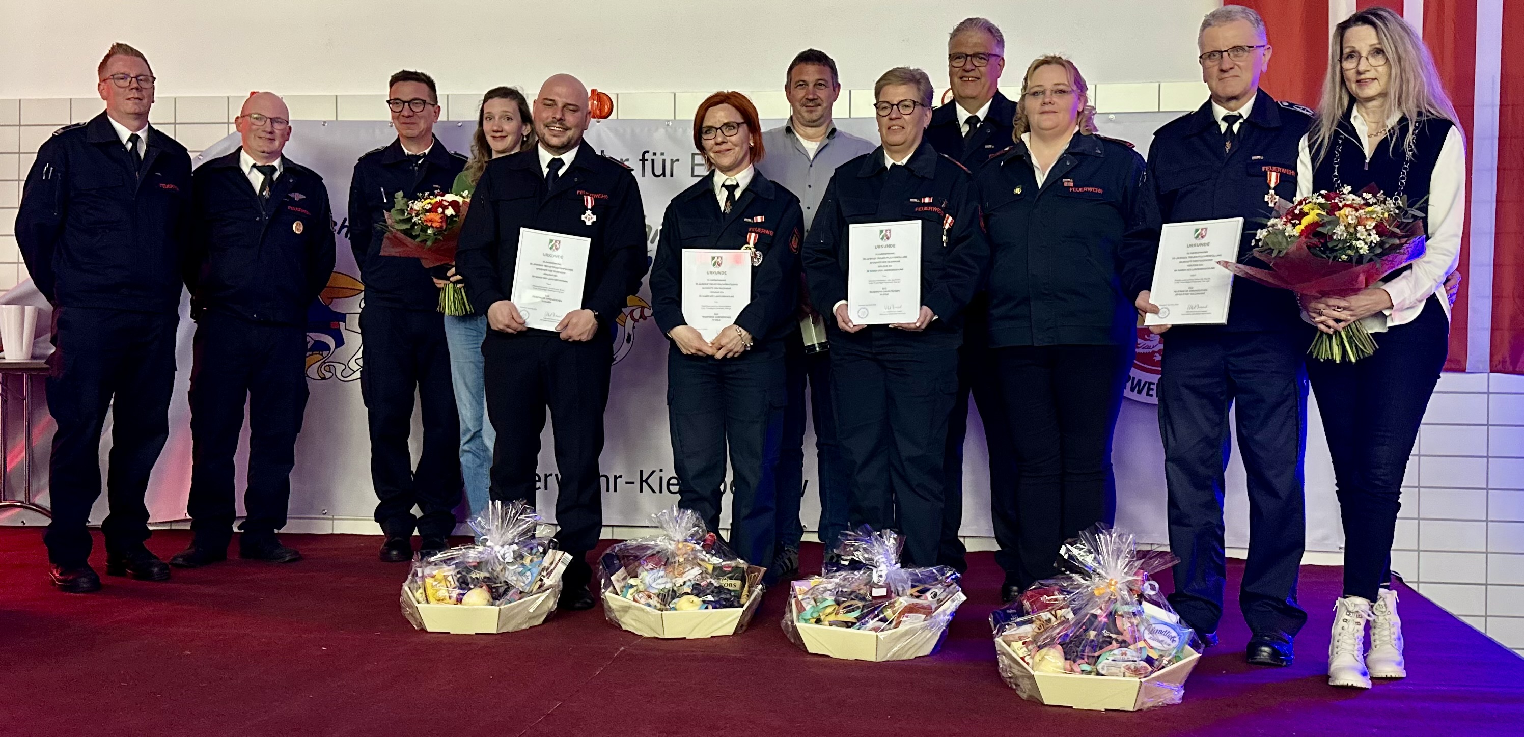 Acht Feuerwehrleute in Uniform stehen nebeneinander auf einer Bühne und halten Urkunden und Blumensträuße. Vor ihnen stehen Geschenkkörbe.