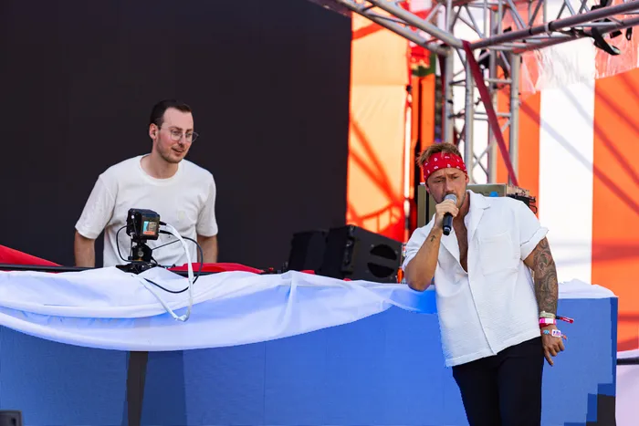 Ein Sänger mit rotem Bandana singt in ein Mikrofon auf einer Bühne beim Ferik Festival, während im Hintergrund ein Mann an einem DJ-Pult steht.