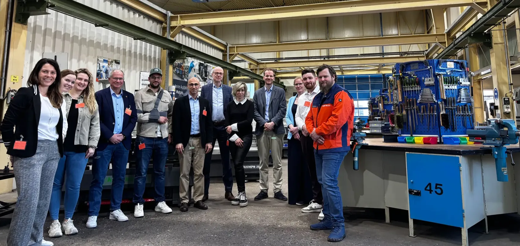 Gruppe von zehn Personen posiert in einer Werkstatt vor einer großen Maschine. Im Hintergrund sind Werkbänke mit Werkzeugen zu sehen.