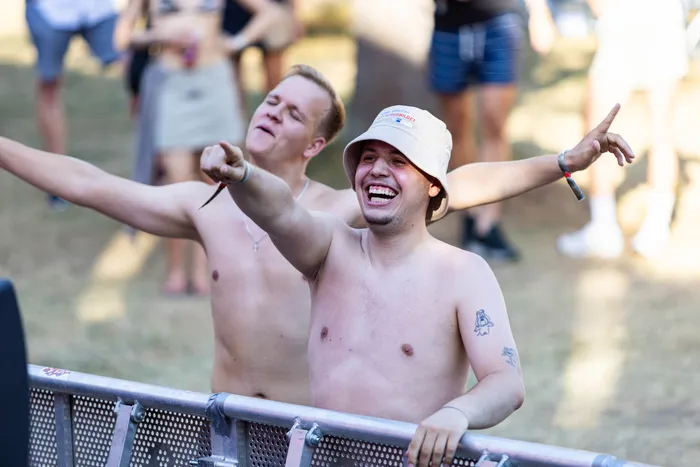 Zwei lachende, oberkörperfreie Männer feiern hinter einer Metallabsperrung, einer davon trägt einen hellen Sonnenhut und streckt die Arme aus.