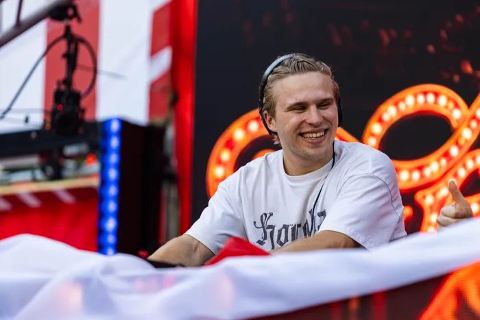 Ein lächelnder Mann mit Kopfhörern und weißem T-Shirt vor einer hell erleuchteten Bühnenkulisse beim Ferik Festival.