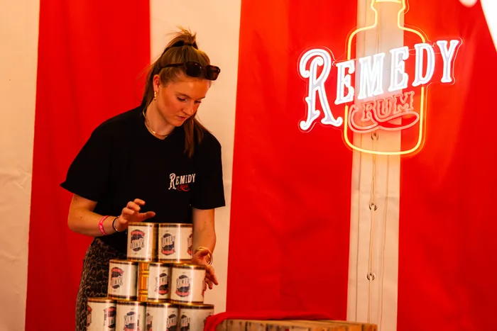 Eine Frau stapelt Dosen von Remedy Rum vor einem rot-weiß gestreiften Hintergrund mit einem leuchtenden Neon-Logo.