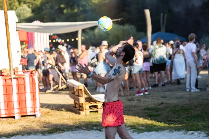 Ein tätowierter, oberkörperfreier Mann spielt mit einem blau-gelben Volleyball im Sand vor einer Menschenmenge auf dem Ferik Festival.