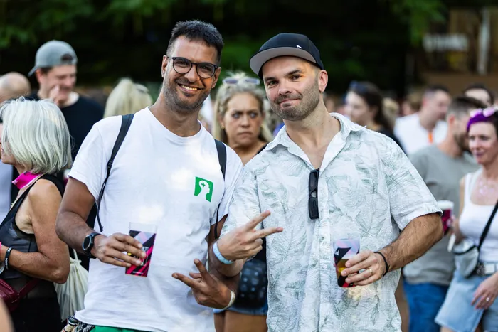 Zwei lächelnde Männer mit Getränken in der Hand posieren vor einer Menschenmenge beim Ferik Festival im Klutertbad.