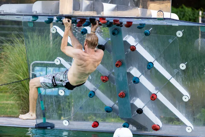 Ein Mann in Badehose klettert an einer transparenten Kletterwand mit bunten Griffen über einem Schwimmbecken.