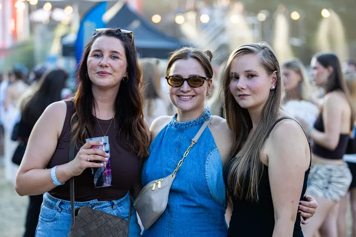 Drei junge Frauen posieren lächelnd beim Ferik Festival vor einer Menschenmenge und Lichtern.