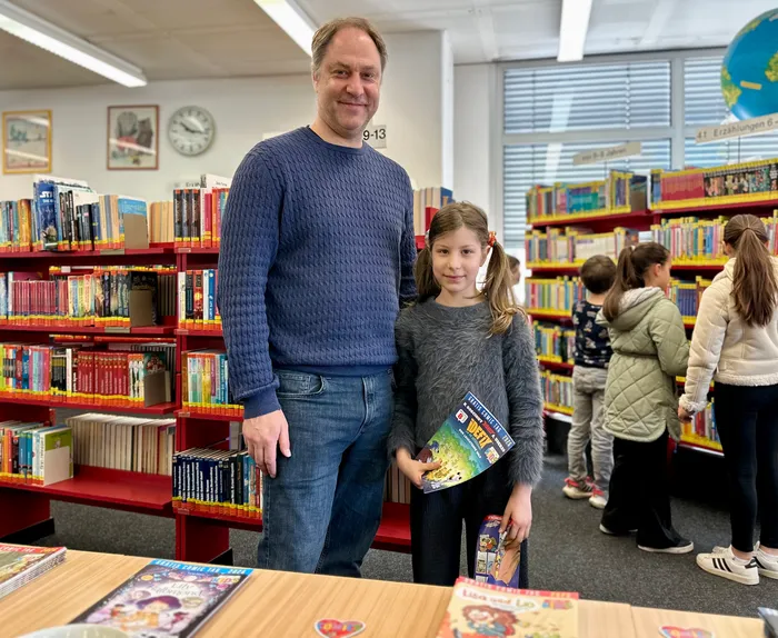 Ein Mann und ein Mädchen stehen vor einem Büchertisch in einer Bibliothek, das Mädchen hält ein Comicbuch in der Hand. Im Hintergrund sind Regale mit Büchern und weitere Personen zu sehen.