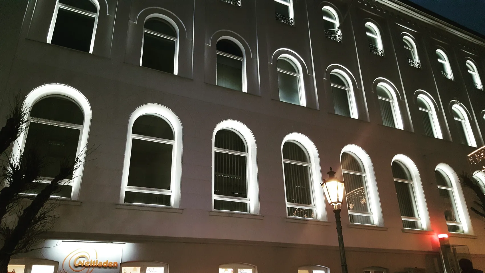 Abendliche Ansicht des VHS-Gebäudes an der Wilhelmstraße mit hell erleuchteten, hohen Fensterbögen und einer historischen Laterne davor.