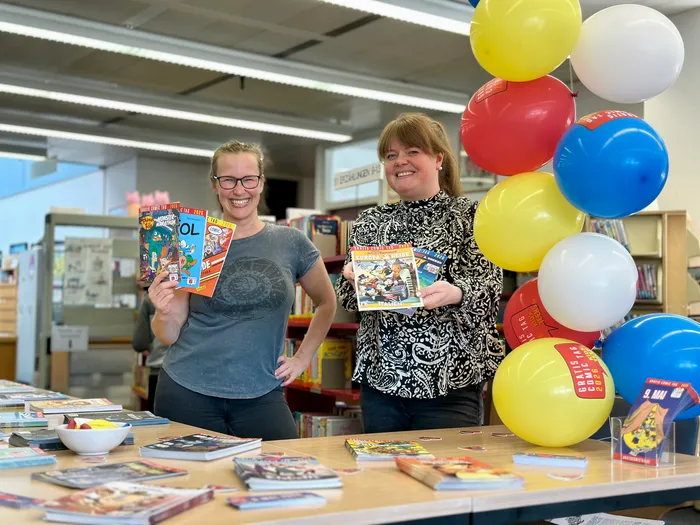 Zwei Frauen stehen in einer Bibliothek und halten jeweils einen Comic in der Hand. Im Hintergrund sind Regale mit Büchern und bunte Luftballons zu sehen.