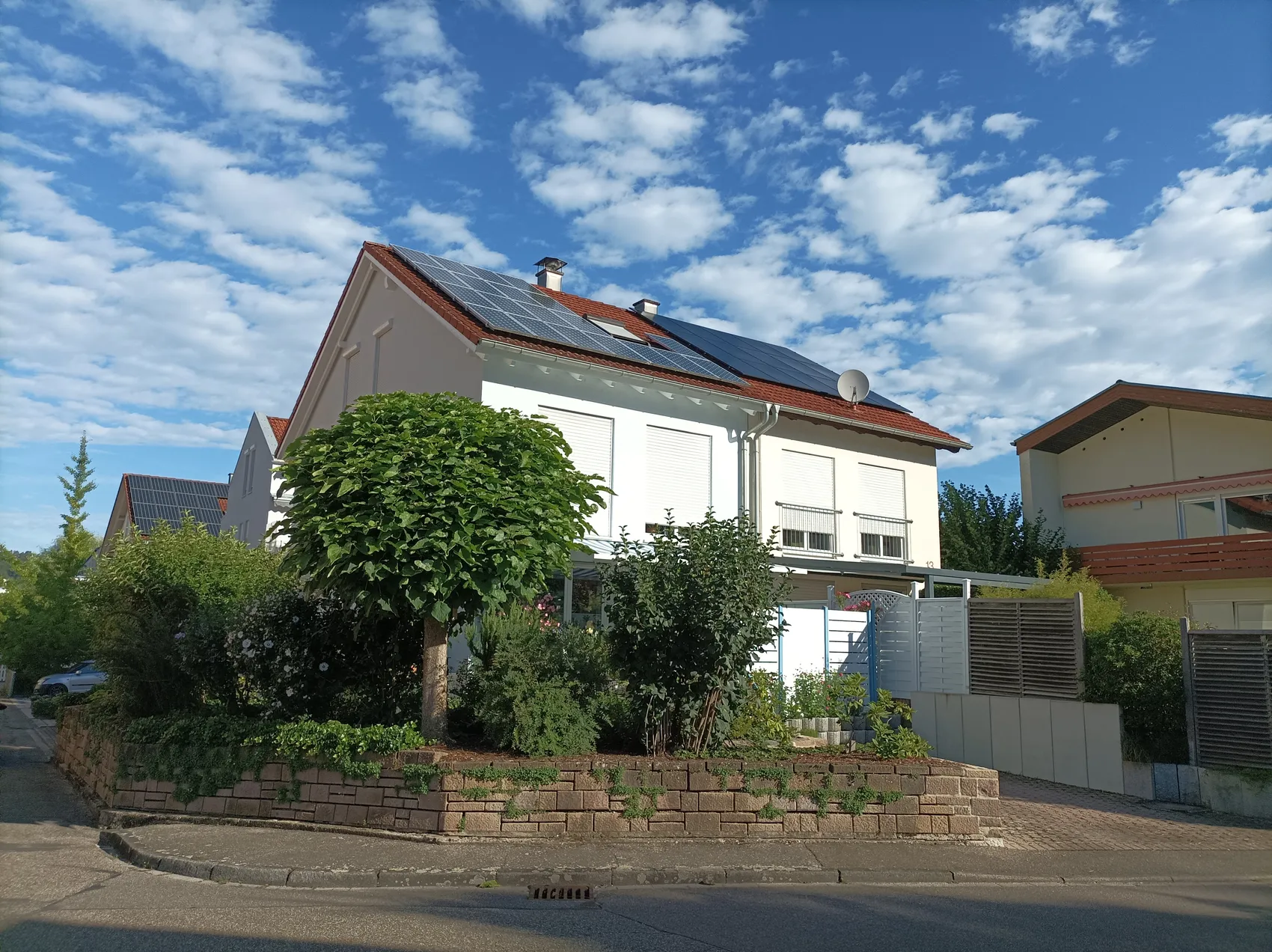 Ein zweigeschossiges Wohnhaus mit Solaranlage auf dem Dach steht im Fokus. Vor dem Haus befindet sich ein gepflegter Garten mit Blumen und Büschen, begrenzt durch eine niedrige Mauer.