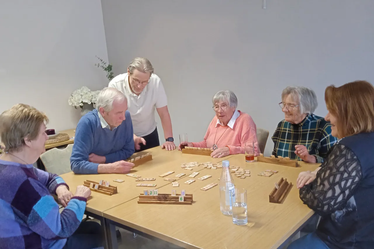Sechs Seniorinnen und Senioren sitzen an einem Tisch und spielen Rommé. Eine Person hilft bei der Spielvorbereitung.