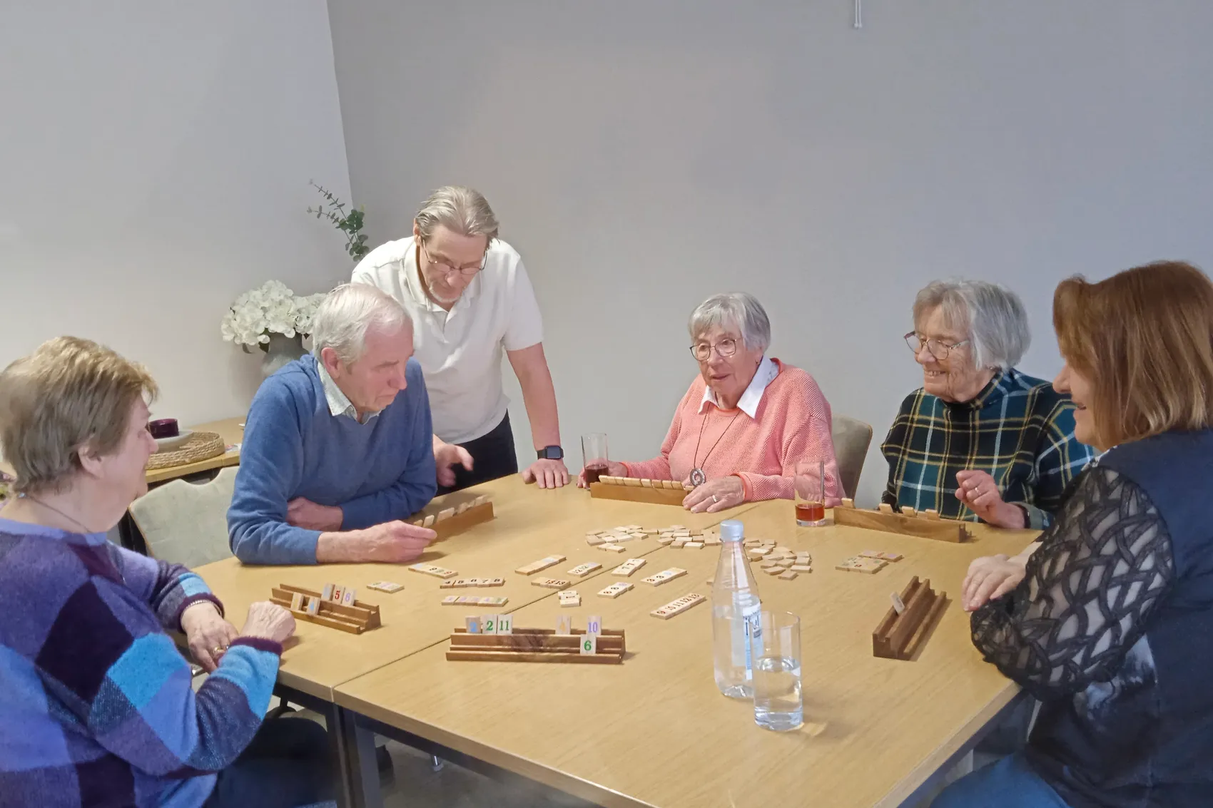 Sechs Seniorinnen und Senioren sitzen an einem Tisch und spielen Rommé. Eine Person hilft bei der Spielvorbereitung.