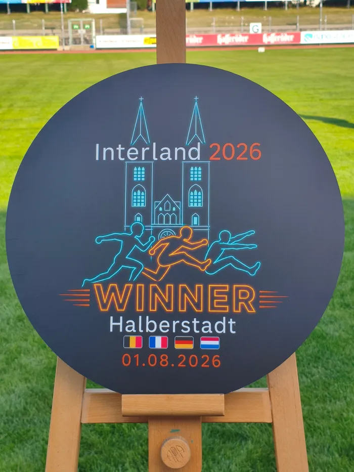 Eine kreisrunde schwarze Tafel auf einem Holzständer mit der Aufschrift „Interland 2026 WINNER Halberstadt“ sowie Grafiken von Läufern und einer Kirche.