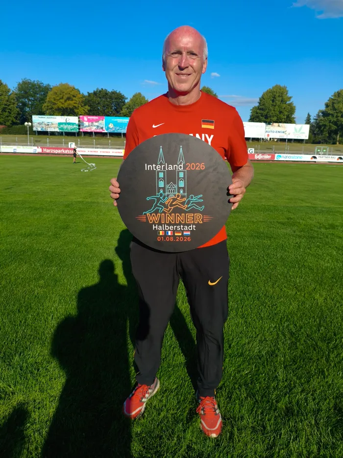 Dirk Maximowitz hält eine große, kreisrunde, dunkle Trophäe mit der Aufschrift „Interland 2026 Winner Halberstadt“ auf einem Sportplatz.