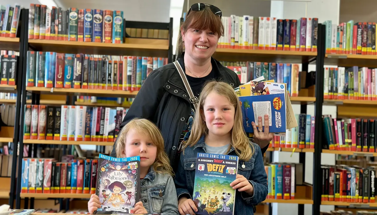 Frau mit zwei Mädchen hält Comicbücher vor Bücherregalen in der Bibliothek.