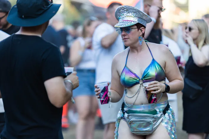 Eine Frau in einem glitzernden Bikini-Oberteil und einer silbernen Kappe hält Getränke beim Ferik Festival.