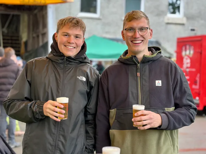 Zwei junge Männer lächeln in die Kamera und halten jeweils ein Glas Bier in der Hand. Im Hintergrund sind weitere Menschen und ein roter Verkaufsstand zu sehen.