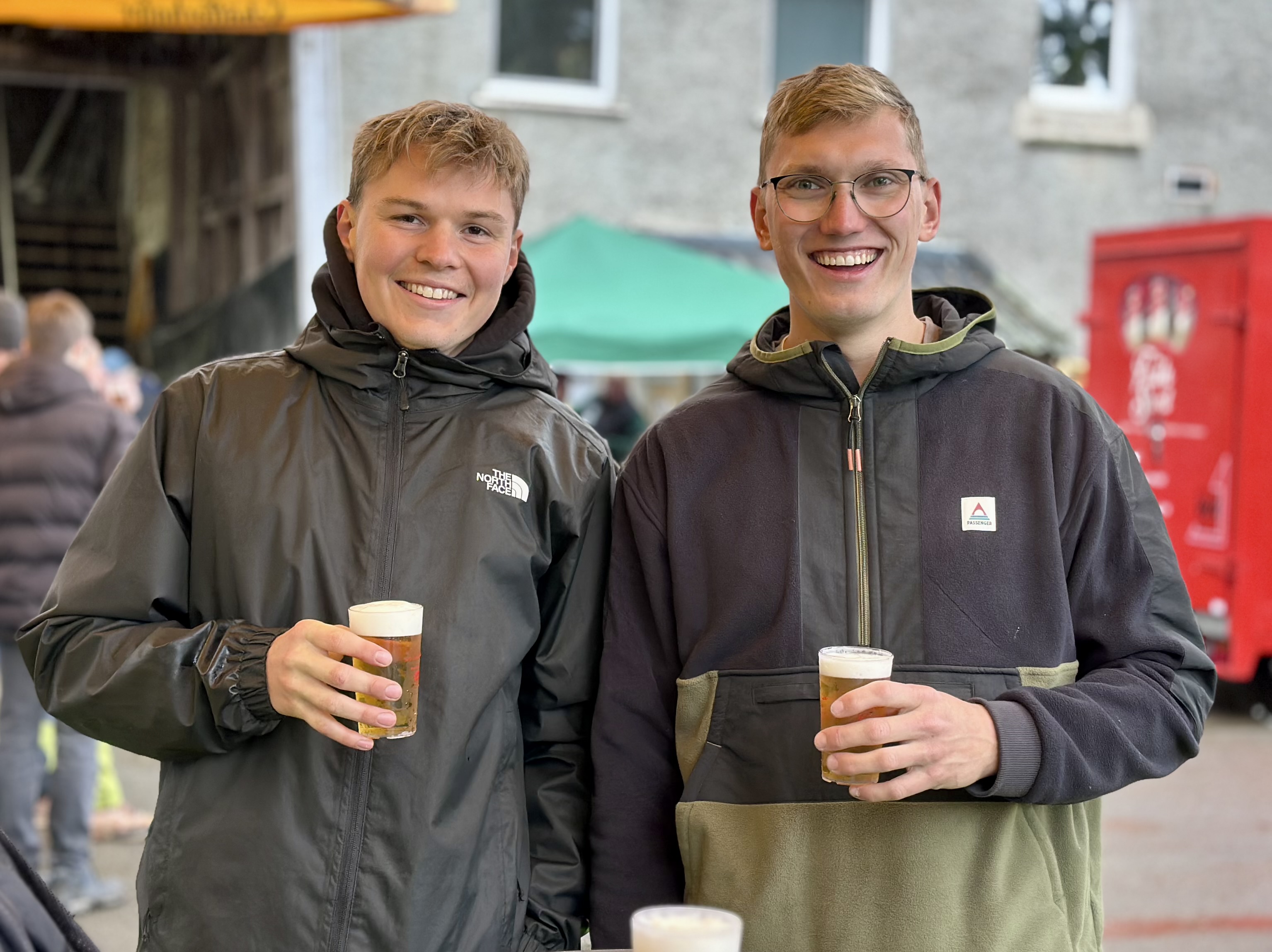 Zwei junge Männer lächeln in die Kamera und halten jeweils ein Glas Bier in der Hand. Im Hintergrund sind weitere Menschen und ein roter Verkaufsstand zu sehen.