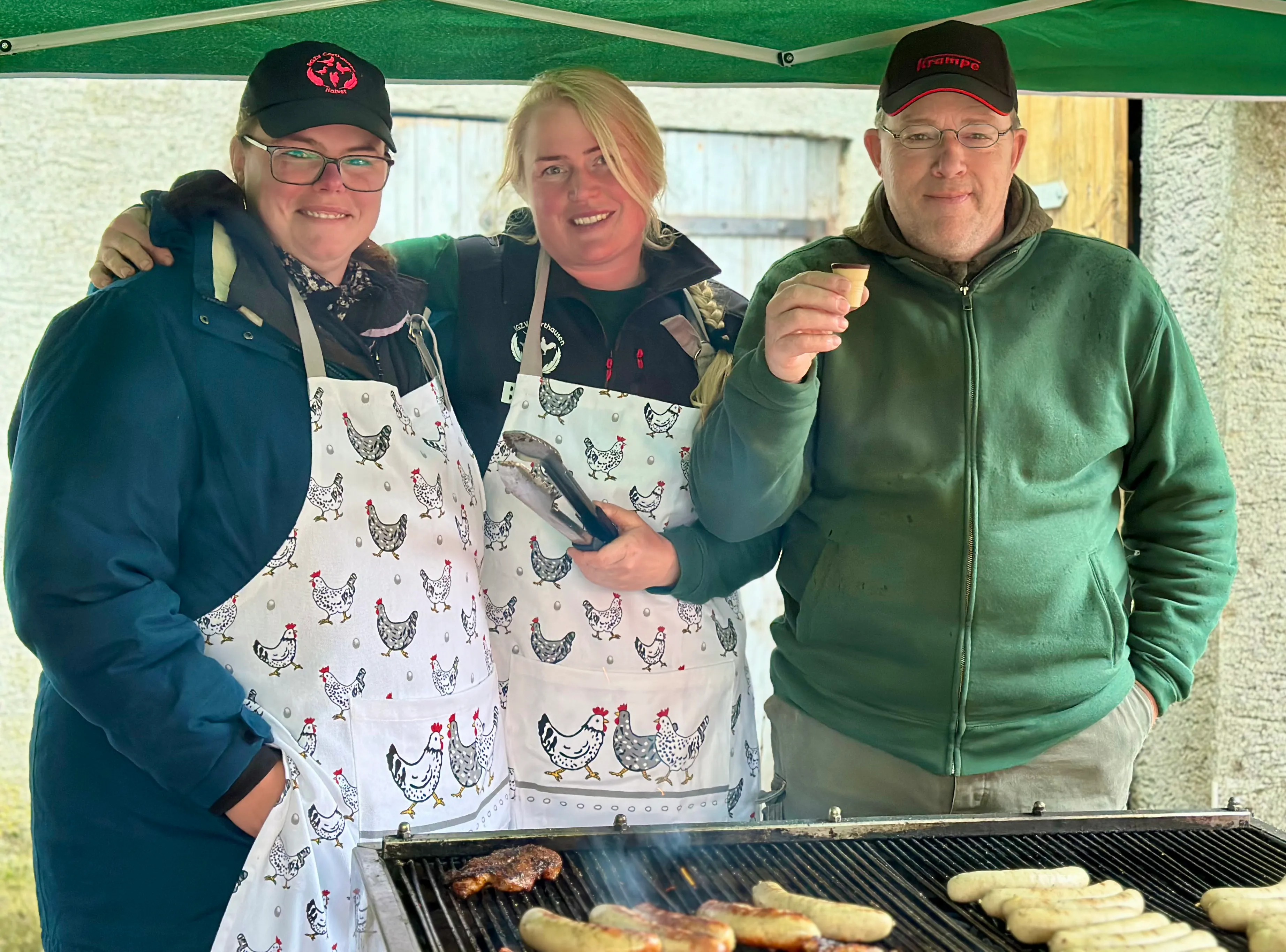 Drei Personen stehen lächelnd hinter einem Grill, auf dem Fleisch zubereitet wird. Sie tragen Schürzen mit Hühnermotiv und Kappen.