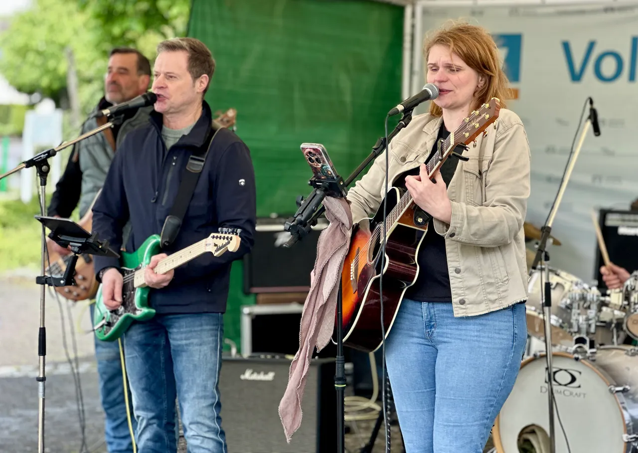 Eine Frau spielt Gitarre und singt in ein Mikrofon, während ein Mann im Hintergrund mit einem E-Bass spielt. Im Hintergrund ist ein Teil einer Bühne und ein Zelt zu sehen.