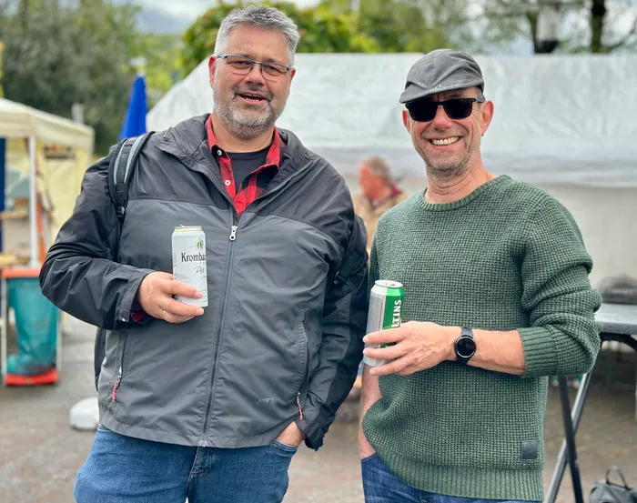 Zwei lächelnde Männer halten jeweils eine Bierflasche in der Hand, im Hintergrund ein Festgelände.