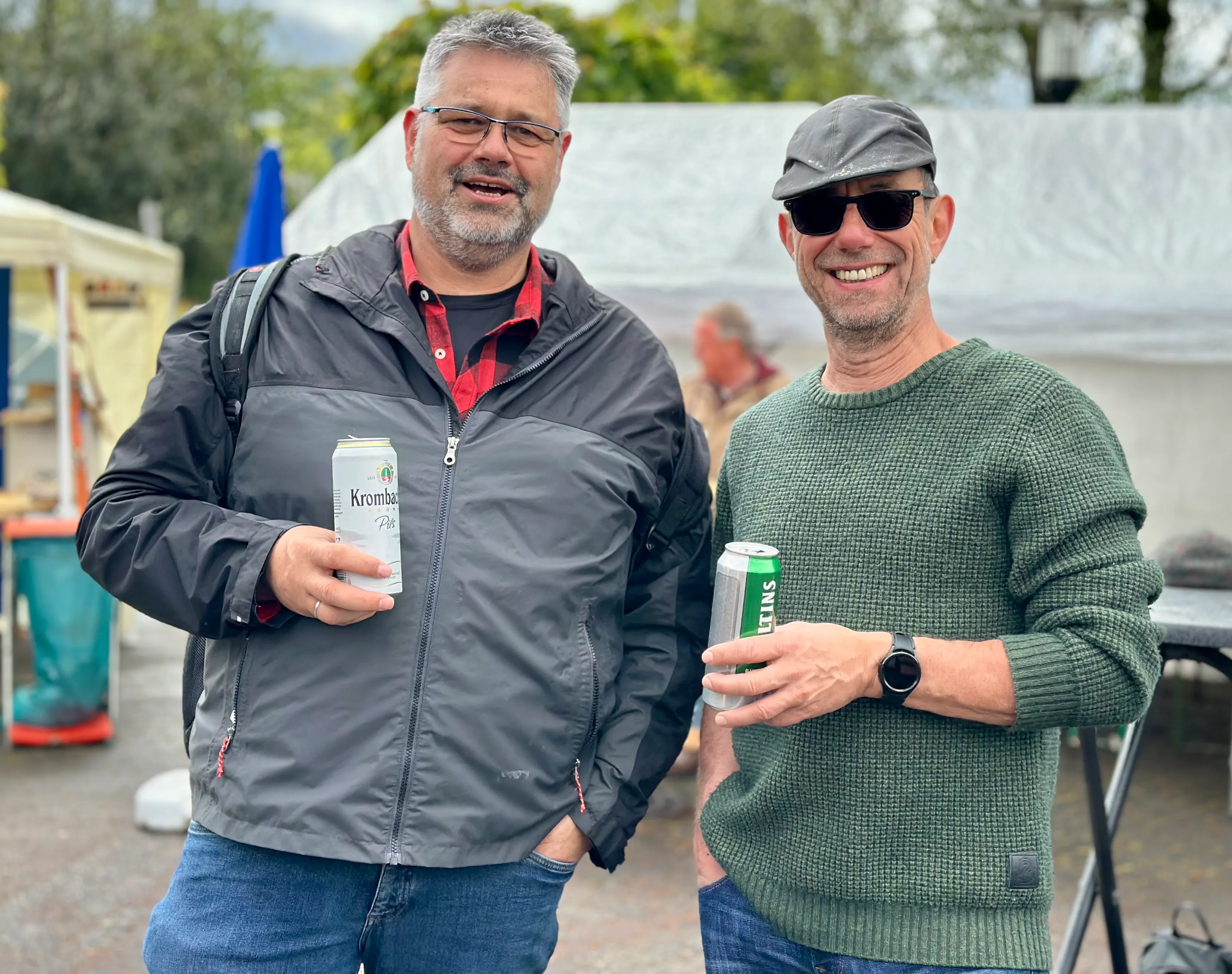 Zwei lächelnde Männer halten jeweils eine Bierflasche in der Hand, im Hintergrund ein Festgelände.