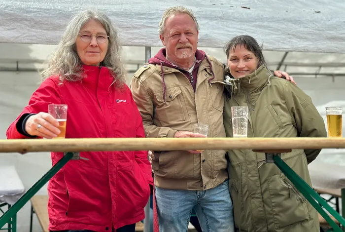 Drei Personen, eine Frau in roter Jacke, ein Mann mit Schnurrbart und eine Frau in olivgrüner Jacke, stehen lächelnd zusammen und halten jeweils ein Glas Bier in der Hand.