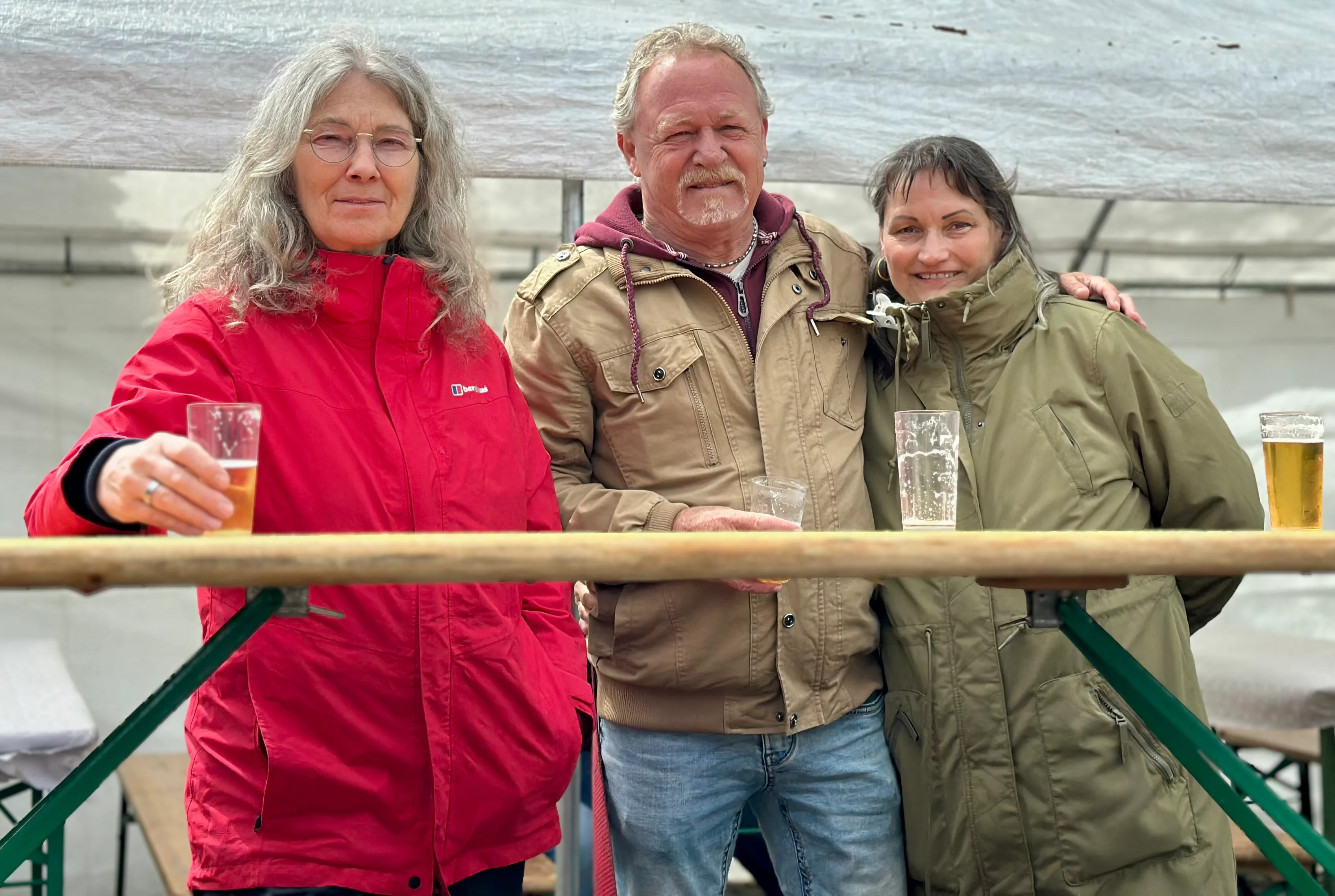 Drei Personen, eine Frau in roter Jacke, ein Mann mit Schnurrbart und eine Frau in olivgrüner Jacke, stehen lächelnd zusammen und halten jeweils ein Glas Bier in der Hand.