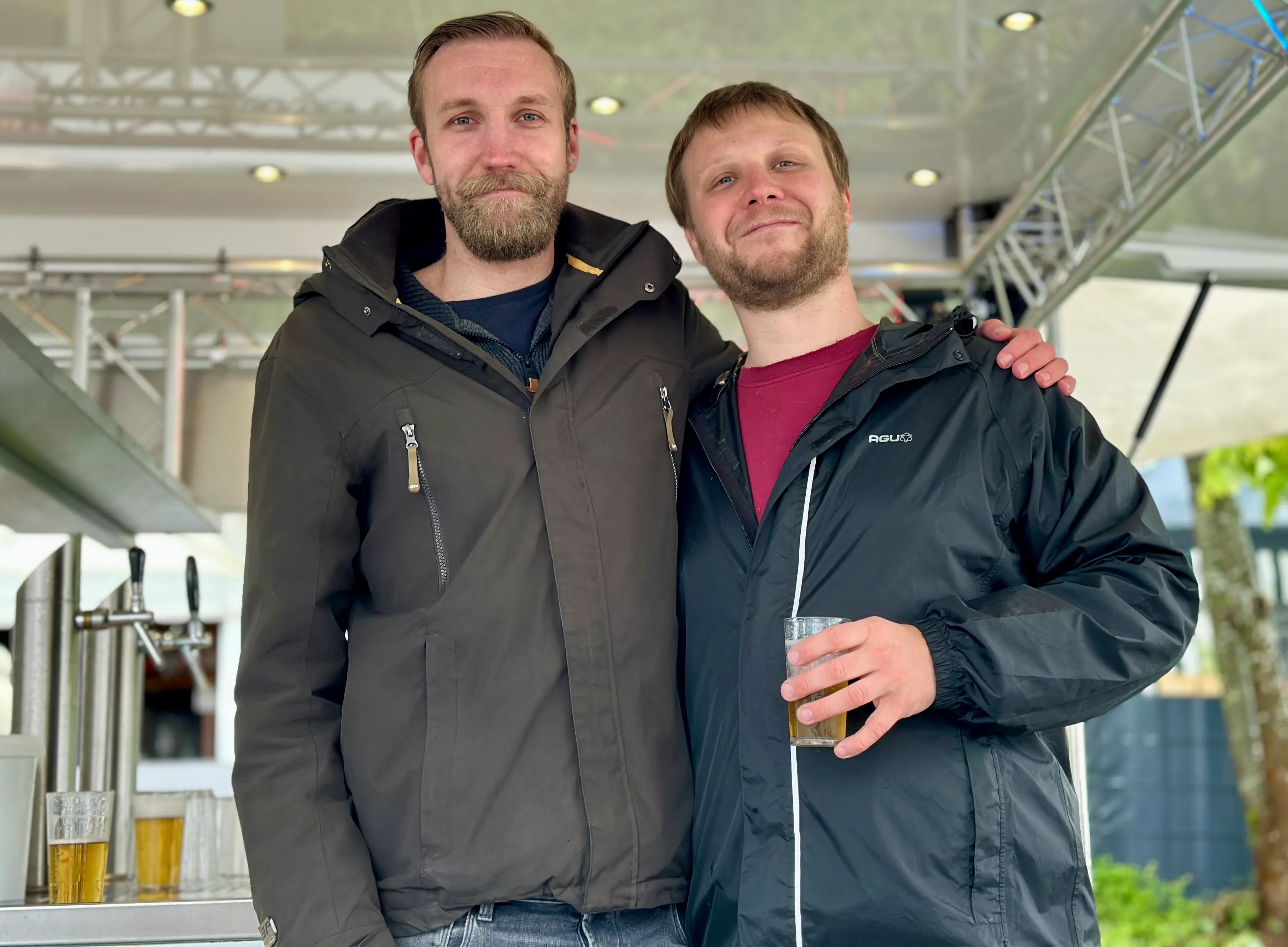 Zwei Männer mit Bart stehen lächelnd vor einer Zapfanlage, einer hält ein Bierglas in der Hand. Im Hintergrund ist ein Zelt zu sehen.