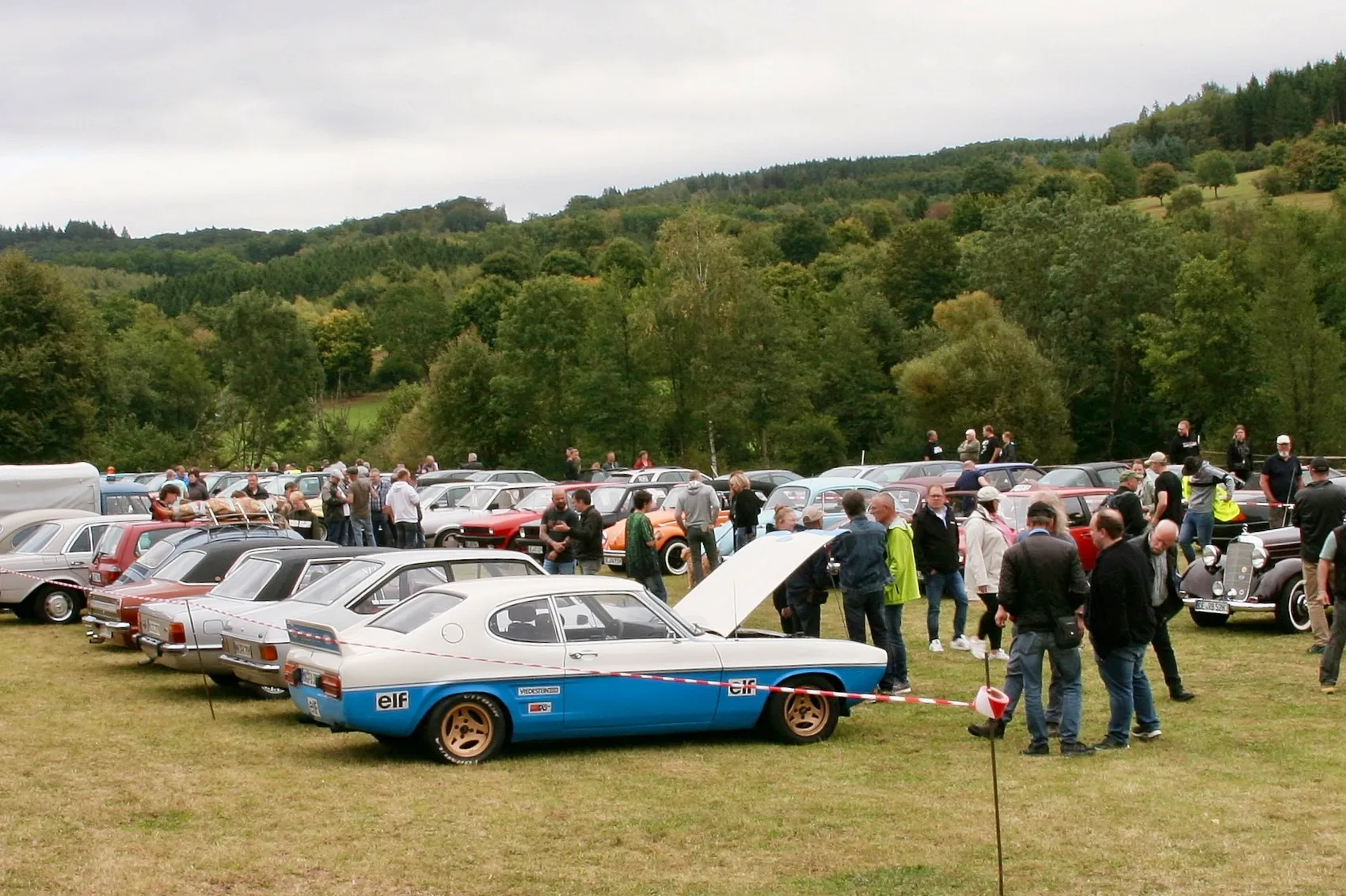 Verschiedene Oldtimer auf einer Wiese bei einem Treffen in Attendorn, im Vordergrund ein blau-weißes Auto mit geöffneter Motorhaube.