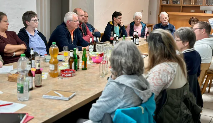 Eine Gruppe von etwa 20 Personen sitzt um einen großen Tisch in einem hellen Raum und nimmt an einer Versammlung teil. Auf dem Tisch stehen Getränke und kleine Snacks.