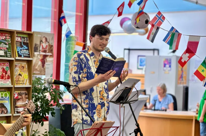 Ein Mann mit Brille liest aus einem Buch vor, während er an einem Mikrofon steht. Im Hintergrund sind Bücherregale, Flaggen und weitere Personen zu sehen.