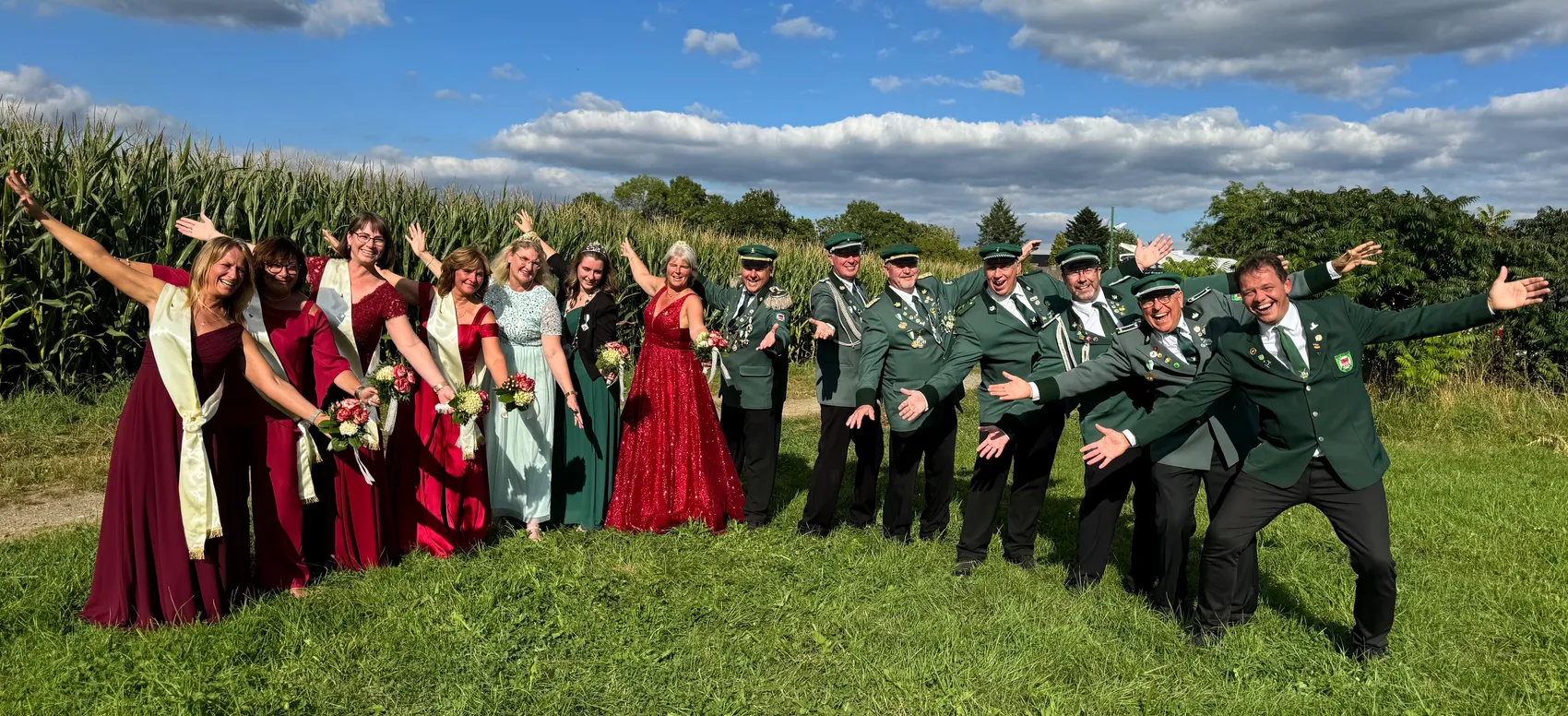 Mitglieder des Schützenvereins in bunten Abendkleidern und grünen Uniformen posieren lachend auf einer Wiese vor einem Maisfeld.