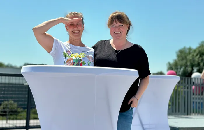 Zwei lächelnde Frauen stehen hinter einem mit weißem Tuch bedeckten Tisch im Freien. Die Frau links hält ihre Hand als Sonnenblende vor die Augen.