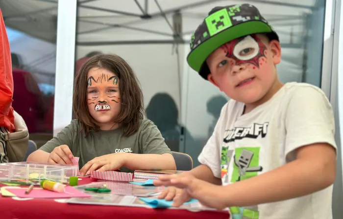 Zwei Kinder sitzen an einem Tisch und basteln. Das Mädchen hat ein Katzen-Gesicht aufgemalt bekommen, der Junge trägt eine Baseballkappe und hat ebenfalls eine Gesichtsbemalung.