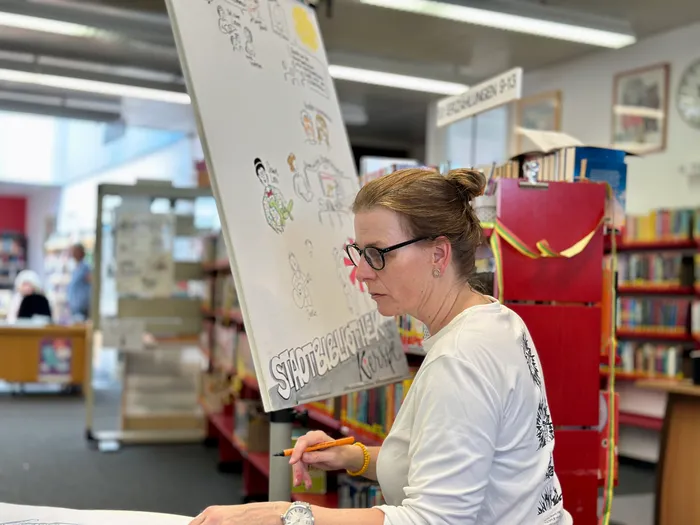 Eine Frau mit Brille und hochgestecktem Haar zeichnet mit einem Stift auf ein großes Flipchart. Im Hintergrund sind Regale mit Büchern und andere Personen in einer Bibliothek zu sehen.
