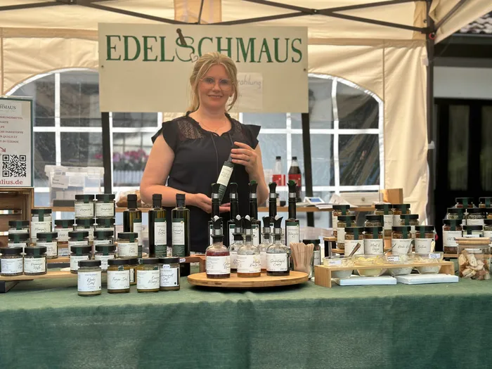 A woman stands behind a table laden with jars and bottles of food products at an outdoor market stall labeled 'EDELSCHMAUS'. The stall is set up under a white tent, and a sign on the left advertises the products available.