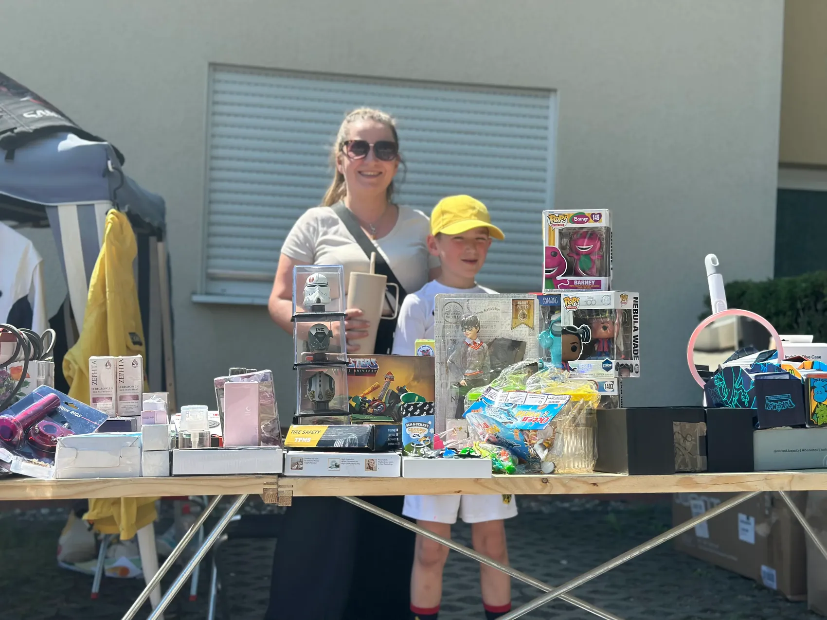 A woman and a child stand behind a table laden with toys, collectibles, and household items at an outdoor flea market. The scene conveys a lively atmosphere with a variety of goods on display.