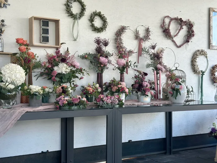 Floral arrangements in varying shades of pink and white are displayed on a long black table draped with a light pink cloth. Behind the table, a white wall is decorated with wreaths and a heart-shaped floral arrangement.