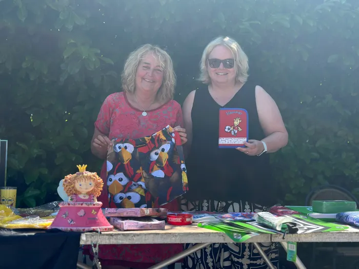 Zwei lächelnde Frauen stehen hinter einem Tisch mit verschiedenen Artikeln, darunter Spielzeug, Bücher und Süßigkeiten, auf einem Flohmarkt im Freien.