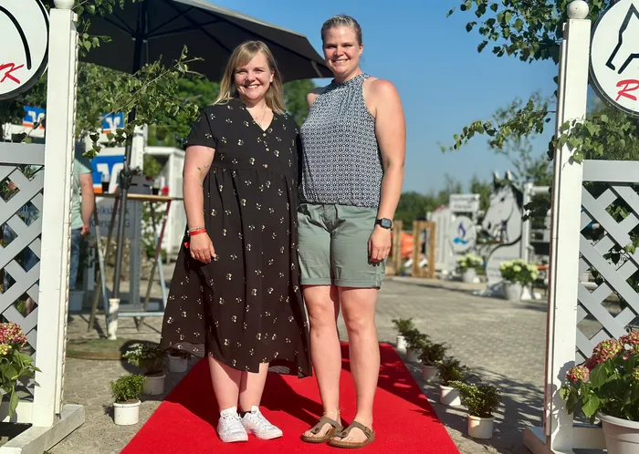 Zwei Frauen stehen lächelnd auf einem roten Teppich vor einem weißen Torbogen mit roten Verzierungen. Im Hintergrund sind weitere Menschen und Pferde zu sehen.