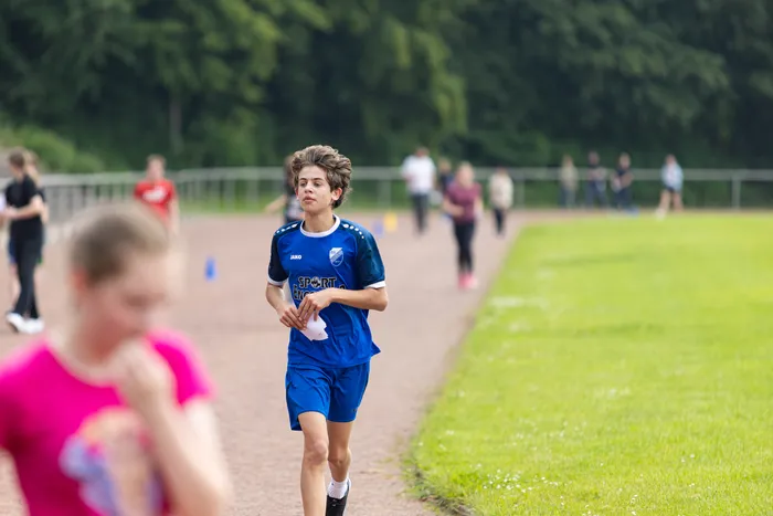 Ein Junge in blauem Sporttrikot läuft während eines Sponsorenlaufs an einer Schule. Im Hintergrund sind weitere Läufer und Zuschauer zu sehen.