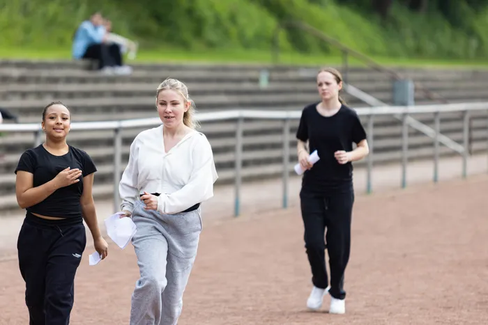 Drei junge Frauen gehen auf einem roten Tartanbahnlaufstrecke. Jede hält mehrere Zettel in der Hand, während sie in Richtung der Kamera blicken. Im Hintergrund sind Zuschauerränge und Bäume zu sehen.