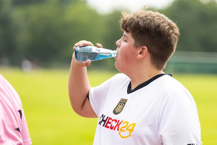 Ein Jugendlicher in einem weißen Fußballtrikot mit dem Logo von CHECK24 trinkt während eines Sponsorenlaufs aus einer Plastikflasche. Im Hintergrund ist ein Fußballfeld zu sehen.